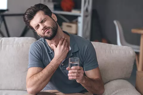 Junger Mann hält sich den Hals und sitzt mit einem Wasserglas auf dem Sofa – Halsschmerzen bei Erkältung